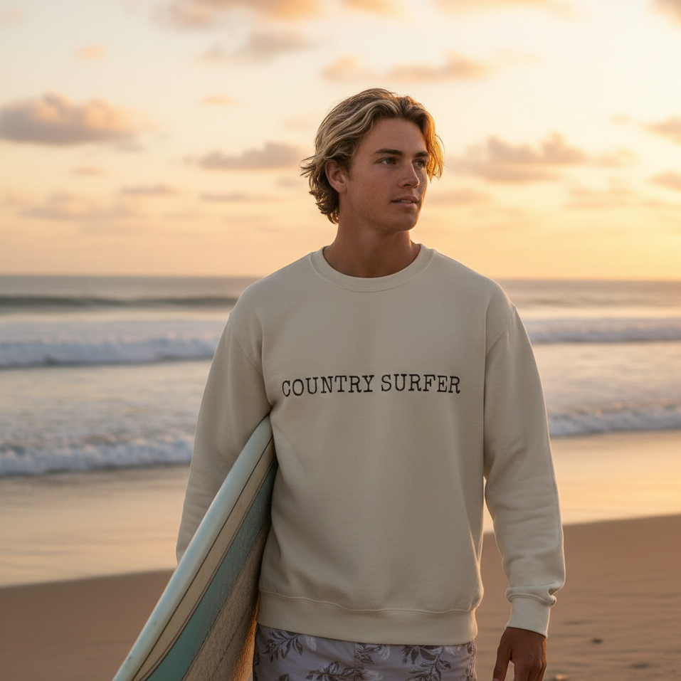 Man holding a surfboard on a beach with 'Country Surfer' sweatshirt at sunset.